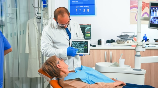 Orthodontiste en gants tenant une tablette et montrant une radiographie de la mâchoire au patient tout en suggérant un traitement pendant la consultation. Stomatologue présentant à une vieille femme une radiographie dentaire à l'aide d'un gadget moderne.