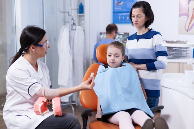 Orthodontiste montrant un échantillon de dents à un enfant et à sa mère au cours d'un examen oral. Petite fille et mère écoutant un stomatologue parlant de l'hygiène dentaire dans une clinique dentaire tenant un modèle de mâchoire