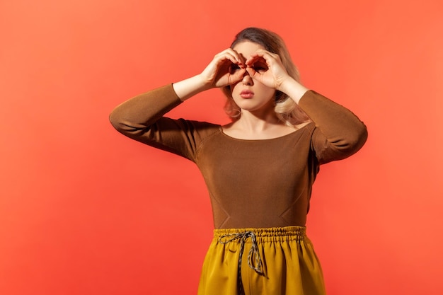 Où vous. Portrait d'une femme blonde sérieuse en chemisier marron décontracté en forme de lunettes, regardant à travers le geste des jumelles. Studio intérieur tourné isolé sur fond rouge.