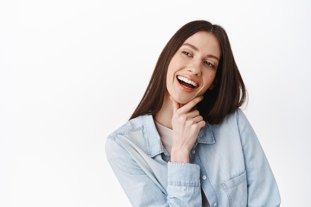 Ouais c'est bien Pensive cool girl touchant le menton souriant et l'air heureux à côté de lire un bon texte promotionnel et acquiescer en signe d'approbation satisfait du produit debout sur fond blanc