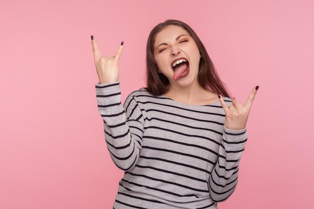 Ouais, génial. Portrait d'une femme à bascule excitée en sweat-shirt rayé montrant un geste de la main rock and roll, des cornes de diable folles avec les doigts et des cris agressifs. studio tourné en intérieur, fond rose