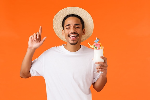 Ouais vacances rocks. Touriste masculin afro-américain joyeux et insouciant s'amusant à boire de l'alcool, tenant un cocktail et faisant la fête à la discothèque de l'hôtel, fond orange