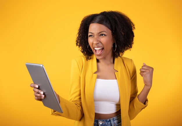 Photo oui dame noire excitée célébrant le succès avec une tablette numérique levant le poing fermé et s'exclamant