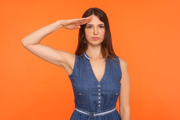 Oui monsieur Femme brune sérieuse attentive en robe denim regardant la caméra avec un geste de salut écoutant l'ordre exprimant la confiance et l'obéissance tourné en studio isolé sur fond orange