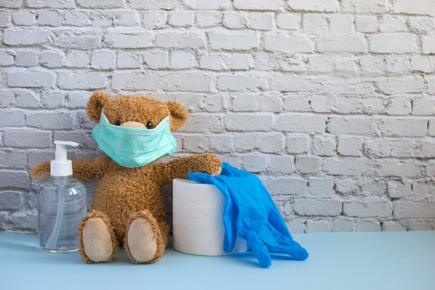 Ours en peluche brun dans un masque médical contenant du gel pour les mains, du papier hygiénique et des gants en caoutchouc bleu.