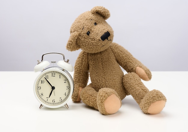 L'ours en peluche brun est assis sur une table blanche et un réveil rond, de cinq minutes à sept heures du matin