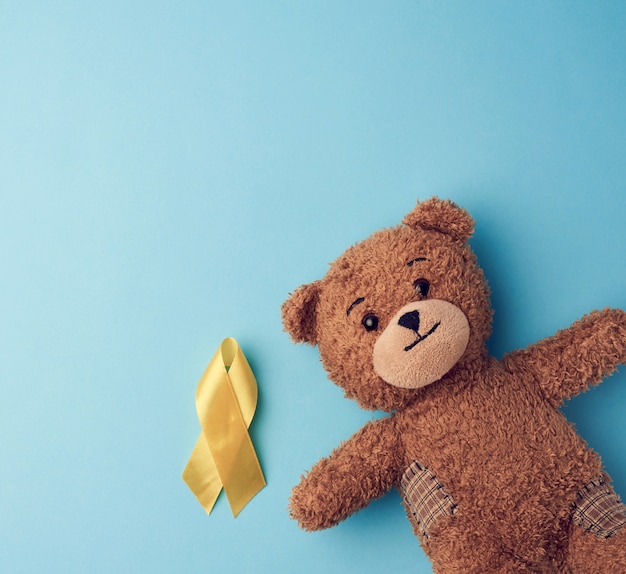 Ours en peluche brun tient dans sa patte un ruban jaune plié en boucle sur un fond bleu. concept de lutte contre le cancer infantile