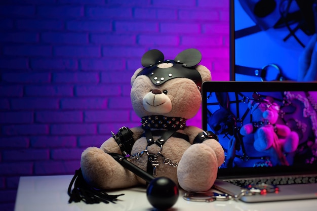 L'ours en peluche dans une ceinture en cuir accessoire pour les jeux BDSM à côté d'un téléviseur portable aux couleurs néon