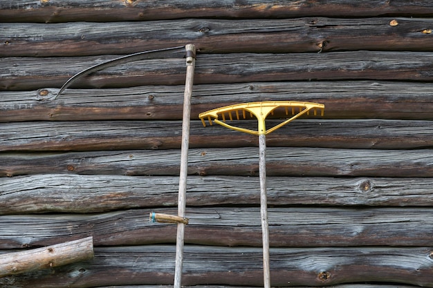Outils agricoles traditionnels faux et rack appuyés contre un mur en bois