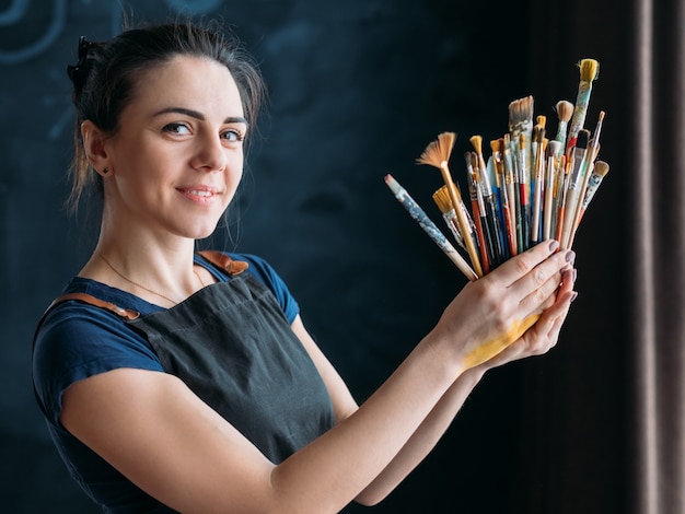 Des outils pour les talents artistiques. Portrait de femme peintre. Belle femme souriante en tablier posant avec des pinceaux.