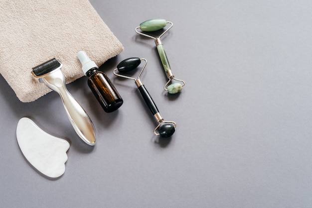 Outils de soin de la peau : rouleau de derma microneedling, rouleaux de massage jade guasha et flacon de sérum sur fond gris