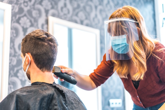 Ouverture de salons de coiffure après la pandémie de coronavirus, covid-19. Mesures de sécurité, masque facial, écran de protection, distance sociale. Salon de coiffure coupe de cheveux avec machine à un garçon client