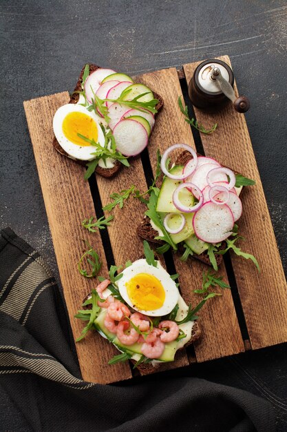 Ouvrez des sandwichs sur du pain de seigle noir avec des œufs, des crevettes, des radis, du concombre, du fromage à la crème et de la roquette pour le petit-déjeuner sur une vieille surface sombre