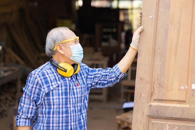 Photo ouvrier âgé, l'artisanat du bois prend sa retraite comme passe-temps pour une bonne retraite, maître professionnel mature masculin asiatique de la fabrication de meubles en bois avec masque protecteur.