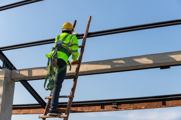 Un ouvrier asiatique de la construction de toit porte un équipement de hauteur de sécurité en montant les escaliers pour installer la charpente du toit, un dispositif antichute pour travailleur avec des crochets pour harnais de sécurité sur le chantier.