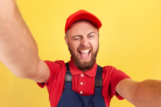 Un ouvrier barbu enjoué portant un uniforme prenant un selfie regardant et faisant un clin d'œil à la caméra POV