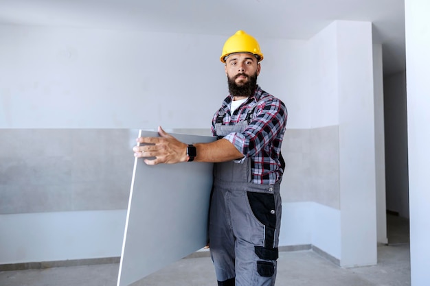 Un ouvrier barbu fort en salopette et avec un casque sur la tête déplace les plaques de plâtre