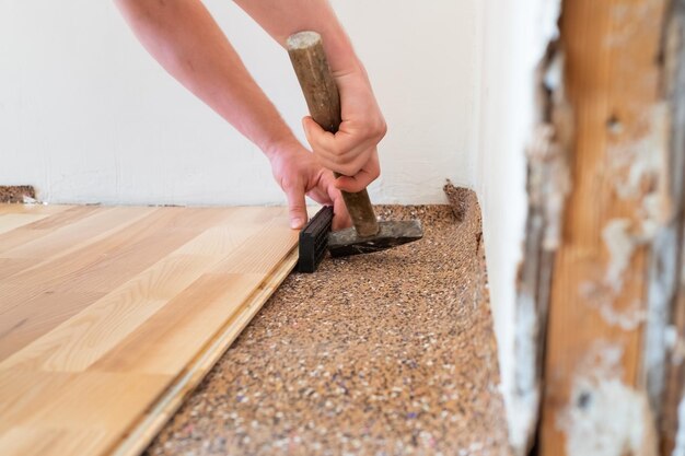 Ouvrier charpentier installant une planche de parquet en bois pendant les travaux de revêtement de sol avec un marteau