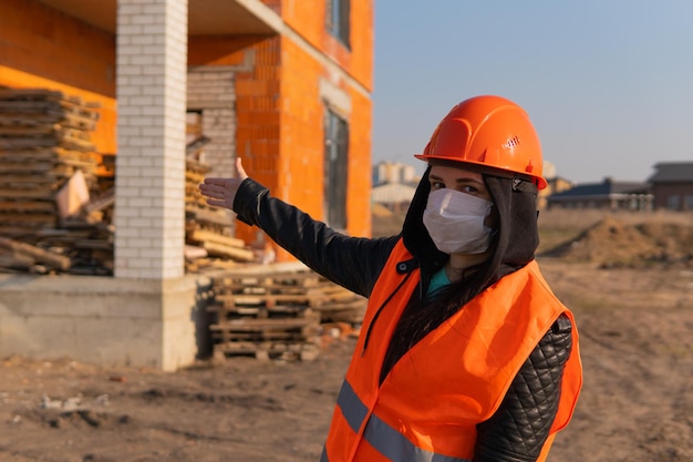 Ouvrier du bâtiment féminin en salopette et en masque médical pointant vers la maison en construction