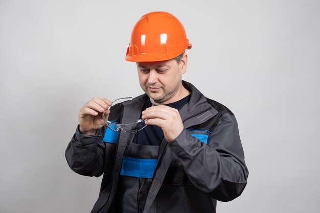 Un ouvrier du bâtiment portant un casque et une combinaison met des lunettes de protection sur un fond blanc