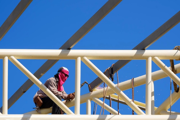 Un ouvrier du bâtiment soude du métal sur la structure du toit du bâtiment sur fond de ciel bleu