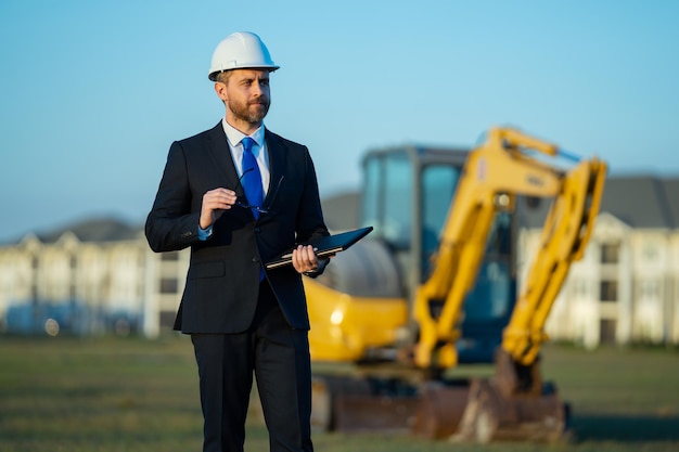 Ouvrier du génie civil sur un chantier de construction Ouvrier du génie mature Homme en costume et casque de protection sur le chantier de construction Ouvrier du génie civil en chef d'âge moyen debout à l'extérieur près de l'excavatrice