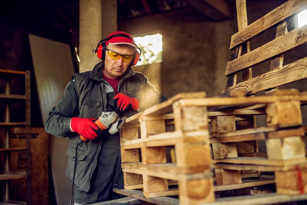 Ouvrier industriel professionnel senior moderne en uniforme avec protection travaillant sur la palette en bois avec le broyeur électrique dans l'atelier.