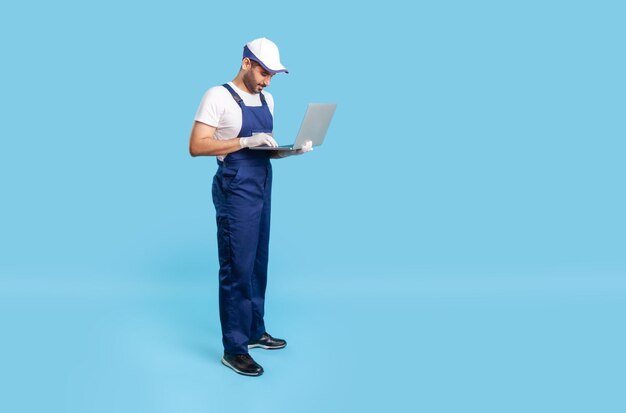 Ouvrier professionnel de pleine longueur en uniforme et gants de protection tapant sur un clavier d'ordinateur portable, ayant une communication en ligne avec le client, lisant la commande pour les services de réparation. tourné en studio intérieur isolé