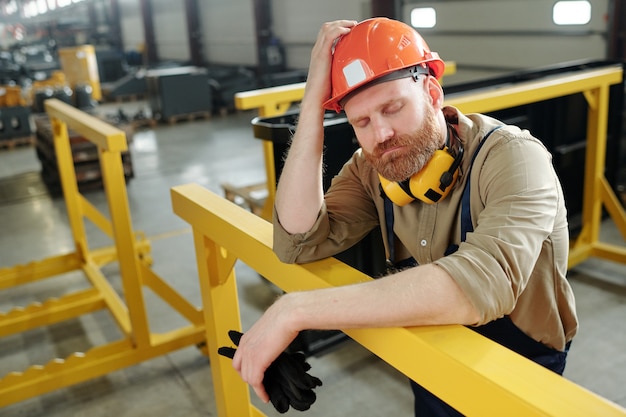 Ouvrier d'usine barbu perplexe gardant les yeux fermés et s'appuyant sur une balustrade en métal jaune