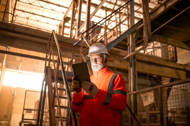 Ouvrier d'usine de l'industrie lourde en uniforme orange haute visibilité et casque tenant une tablette et vérifiant la production.