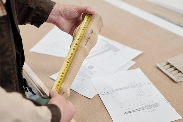 Photo ouvrier d'usine moderne mesurant une partie en bois de meubles tout en le maintenant sur des croquis sur la table et en comparant l'indication de la longueur