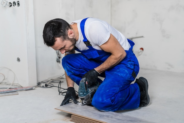 Photo ouvrier en vêtements de travail bleus scier des planches stratifiées avec une scie électrique