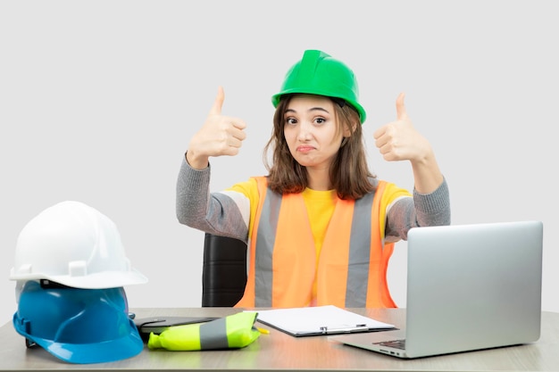 Ouvrière en uniforme assise au bureau et montrant les pouces vers le haut. Photo de haute qualité