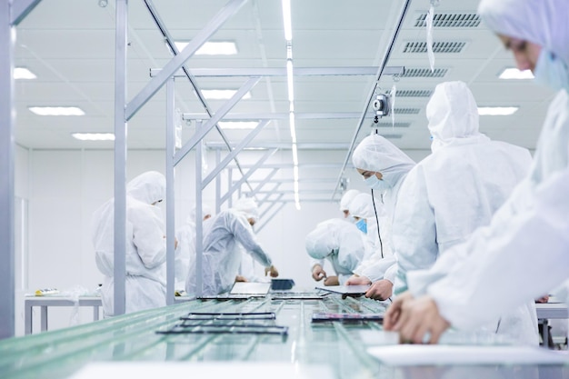 Photo ouvriers d'usine en combinaisons de laboratoire blanches et masques faciaux produisant des téléviseurs sur une chaîne de montage verte avec des équipements modernes