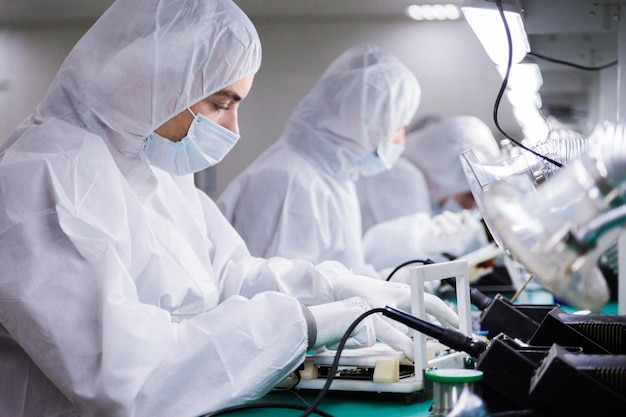 Ouvriers d'usine en combinaisons de laboratoire blanches et masques faciaux produisant des téléviseurs avec des fers à souder sous des lampes lumineuses