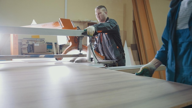 Les ouvriers de l'usine coupent un fragment de bois sur une scie électrique dans l'industrie du meuble, vue large