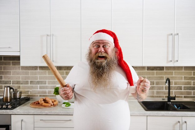 Overweight man in Santa hat tenant un rouleau à pâtisserie en bois sur une main et serre son poing