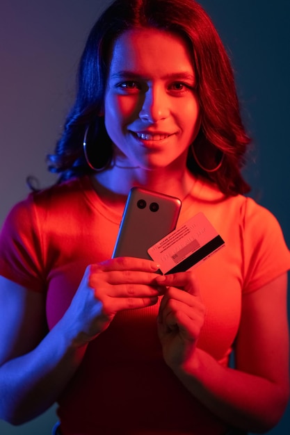 Paiement sécurisé Cyber-bancaire Achats sur Internet Femme souriante joyeuse en lumière de couleur bleu rouge vif néon avec carte de crédit téléphonique avec code CVV sur fond violet foncé