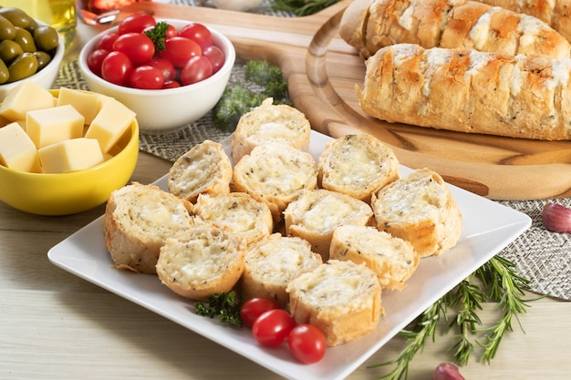 Pain à L'ail Dans Une Assiette Carrée Blanche Sur La Table Avec Du Fromage, Du Romarin, Des Olives Et Des Tomates Cerises.