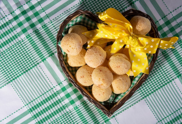 Pain au fromage, panier en forme de coeur avec un noeud de ruban jaune rempli de pain au fromage sur une nappe à carreaux, vue du dessus.