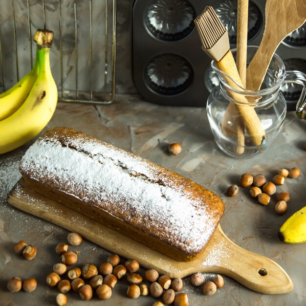 Le pain aux bananes est allongé sur le tableau parmi les ustensiles de cuisine, les bananes et les noisettes