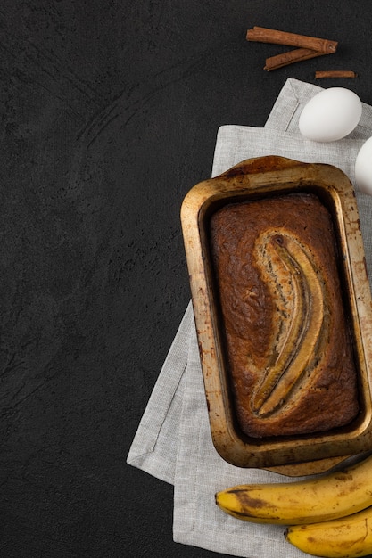 Pain aux bananes frais prêt dans un plat de cuisson rectangulaire avec des ingrédients sur fond sombre