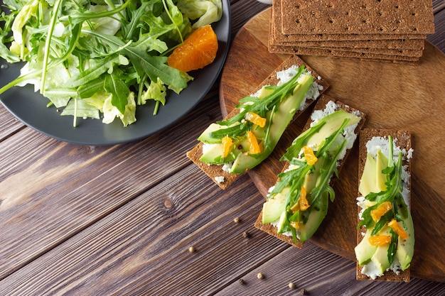 Pain croustillant à grains entiers avec fromage, avocat et feuilles fraîches avec salade sur fond en bois.