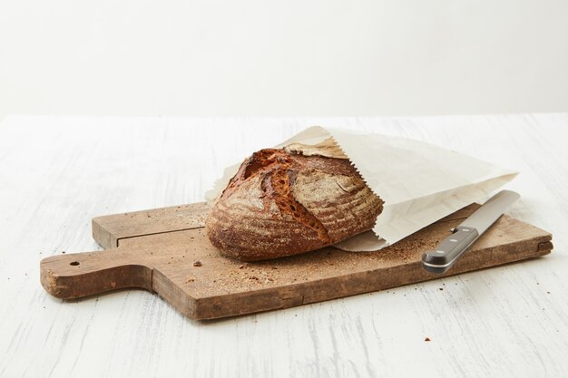 Pain fait maison enveloppé dans du papier sur une planche en bois avec un couteau sur fond clair