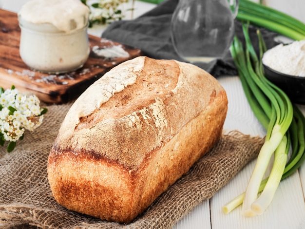 Pain frais sur la toile de jute, le levain et la farine avec une cruche d'eau sur une table en bois blanc