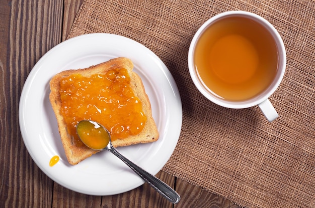 Pain grillé avec de la confiture d'abricot dans une assiette et une tasse de thé noir sur la vue de dessus de table en bois