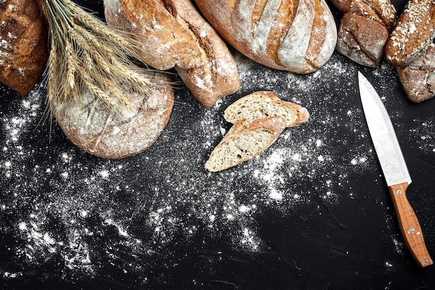 Photo pain de seigle fait maison saupoudré de farine et de divers grains et graines sur fond noir avec des épillets de blé ou de seigle et d'avoine