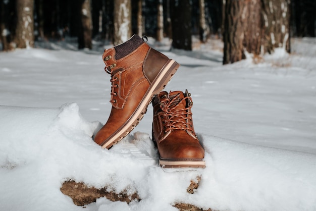 Paire de bottes d'hiver en cuir marron pour hommes debout sur une bûche dans une forêt enneigée aux beaux jours, à l'extérieur. Chaussures de randonnée et de voyage. Objet de vêtement, pas de personnes.