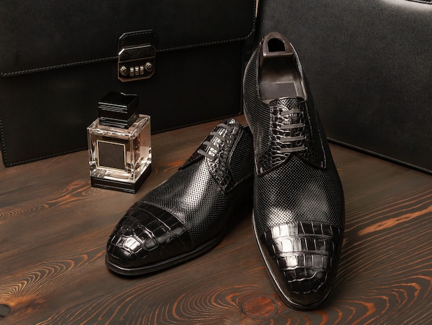 Une paire de chaussures noires pour hommes repose sur une surface de planches sombres et jetées, à côté d'une bouteille de parfum sur fond de deux porte-documents en cuir pour hommes. Vue de côté. Style de patron