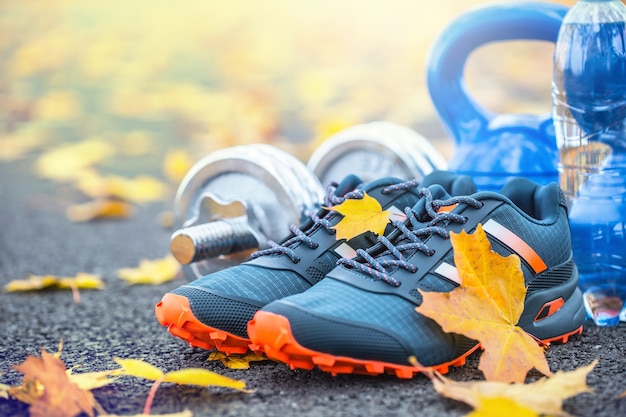 Paire de chaussures de sport bleu eau et haltères posées sur un chemin dans une allée d'automne avec des feuilles d'érable